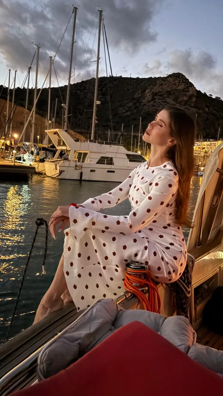 Modelo con vestido blanco sentada en la proa del velero al anochecer, con el puerto y la montaña del Garraf al fondo