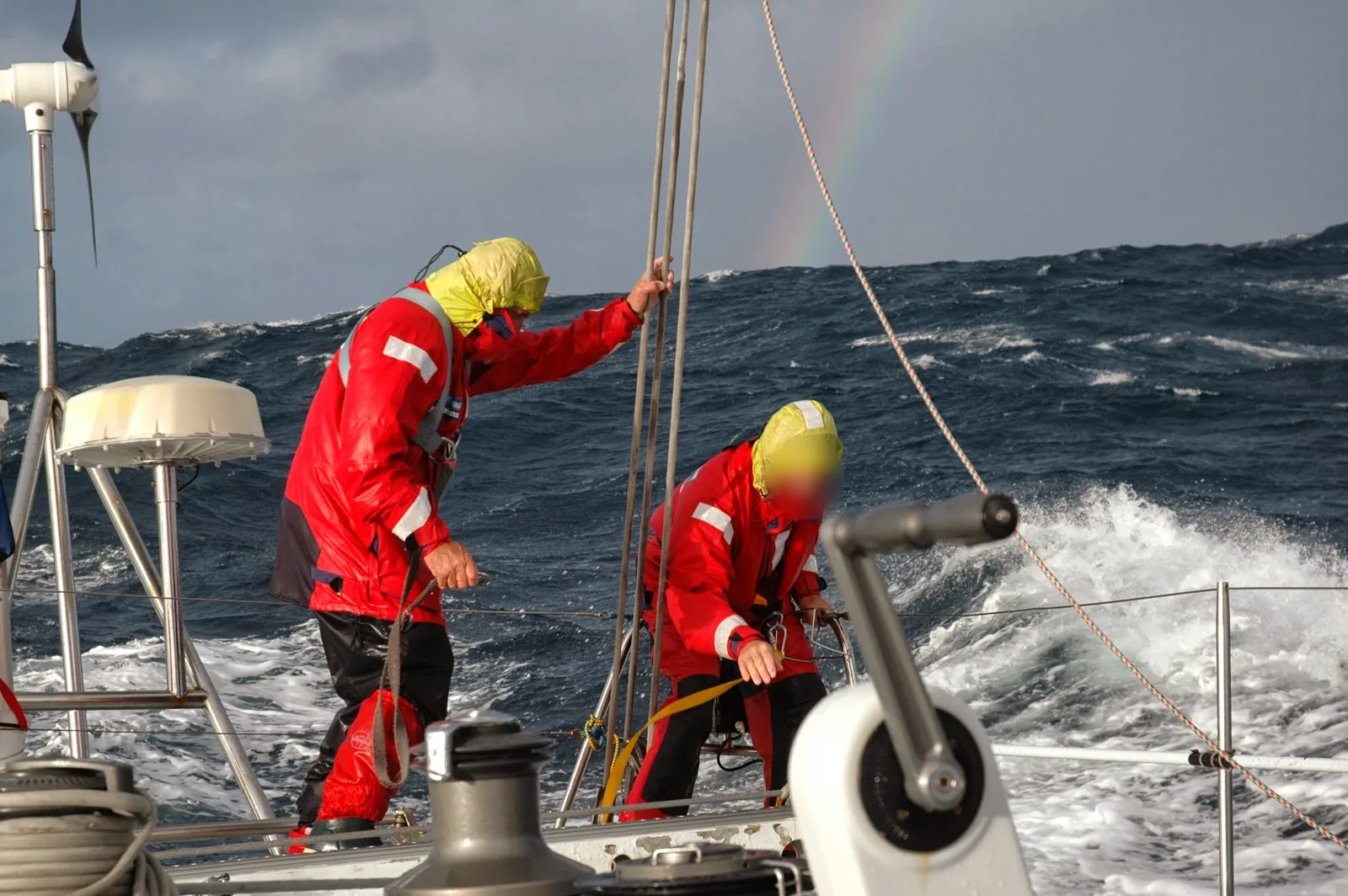 Dos tripulantes con trajes de aguas trabajando en cubierta con mar gruesa y un arco iris al fondo